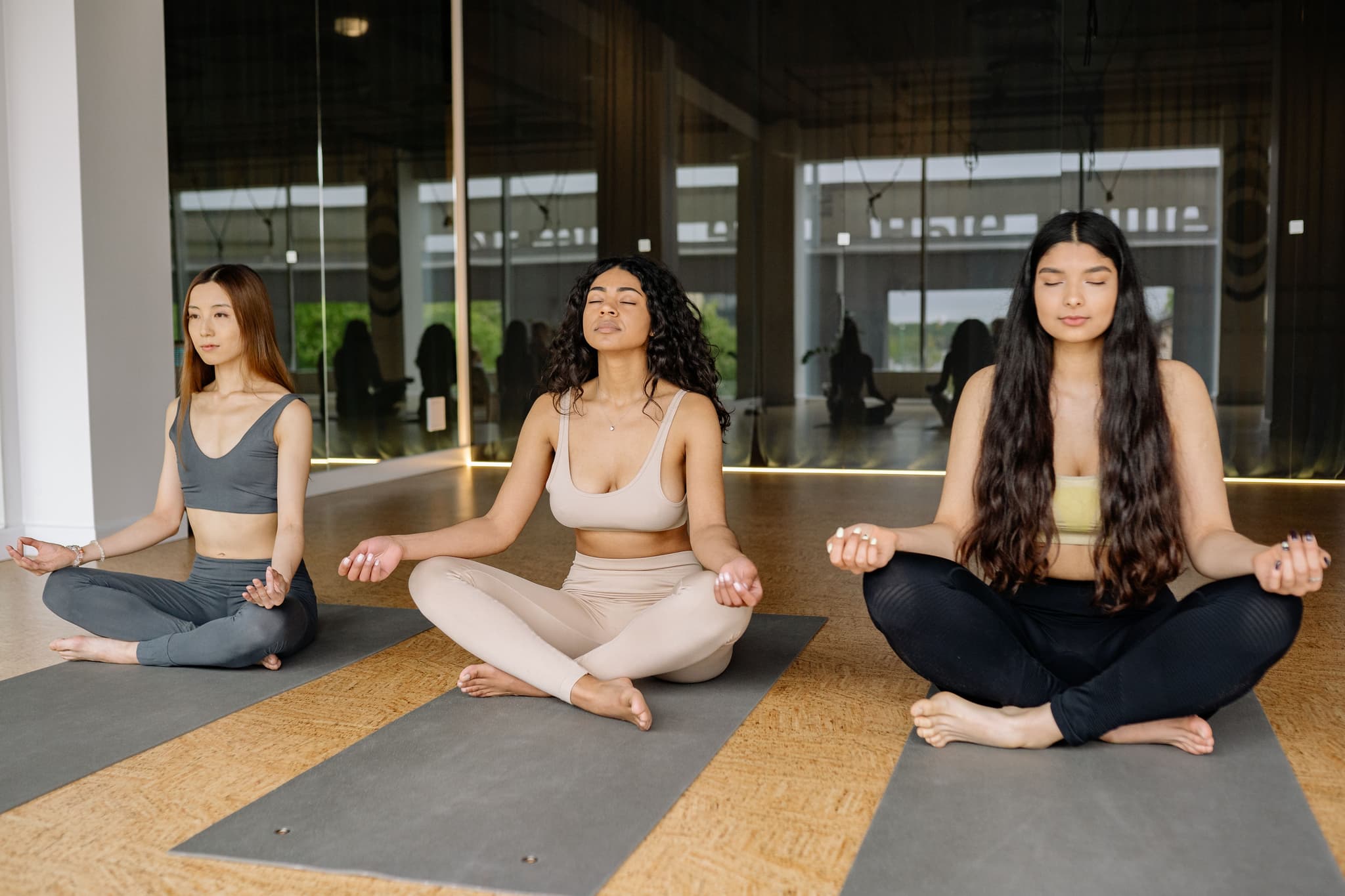 Group Meditation in Yoga Studio Three people sitting in cross-legged meditation pose on yoga mats in a bright studio with floor-to-ceiling windows.