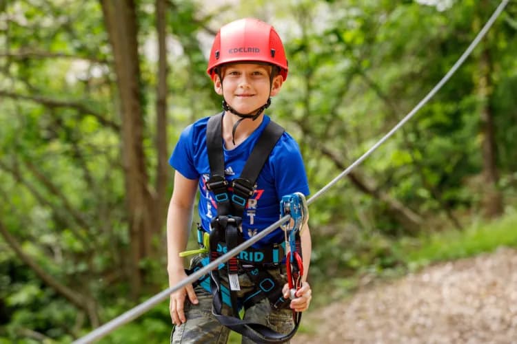 Child on Zipline Through Forest Child in red helmet and blue shirt on a zipline through a forested area, wearing safety harness and gear.