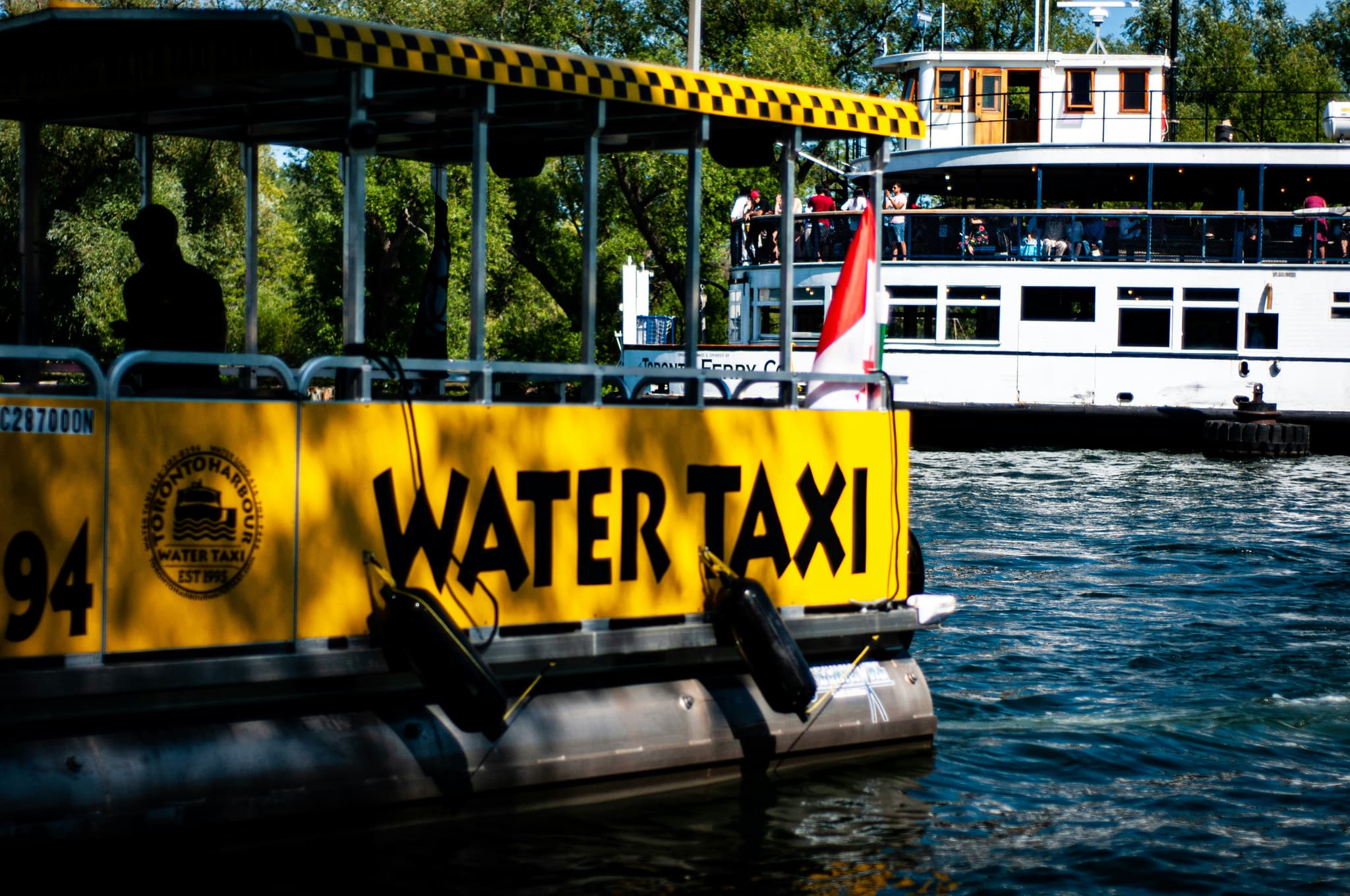 Yellow Water Taxi at Waterfront Dock Yellow water taxi boat with black lettering moored at a waterfront dock, with passenger vessels and trees in the background.