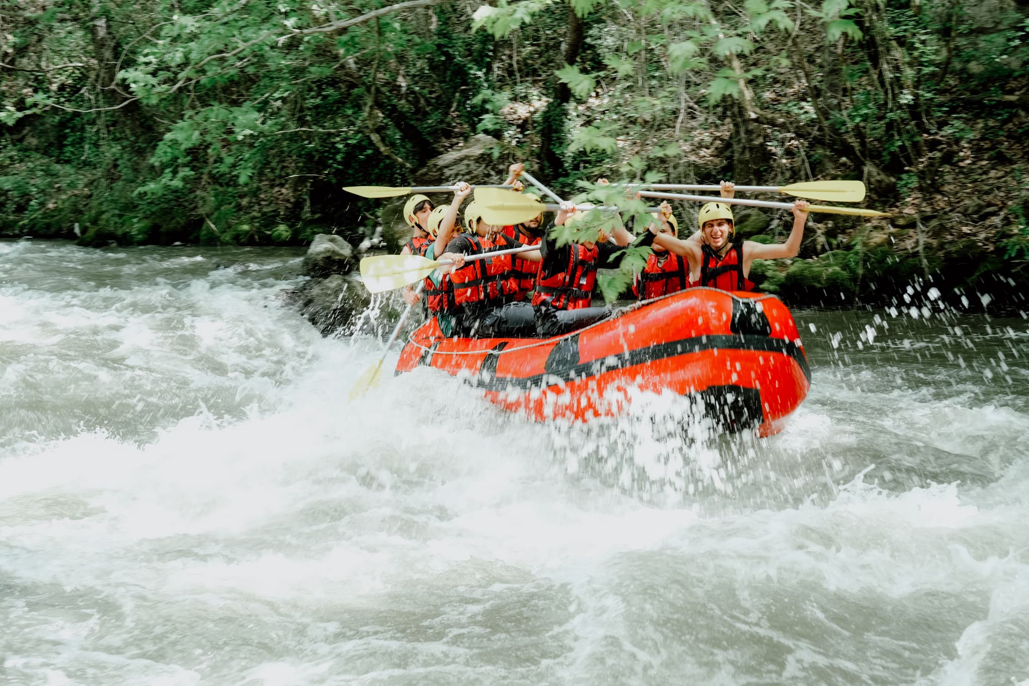 Whitewater Rafting Through River Rapids Group of people whitewater rafting through river rapids in a red inflatable boat, surrounded by dense forest.