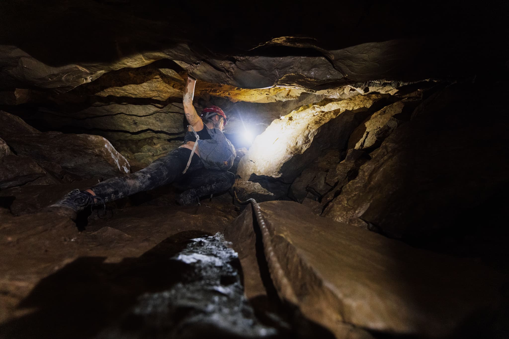 Caver Navigating Underground Cave Passage Caver with headlamp navigating a narrow rocky passage while holding a rope in an underground cave system.