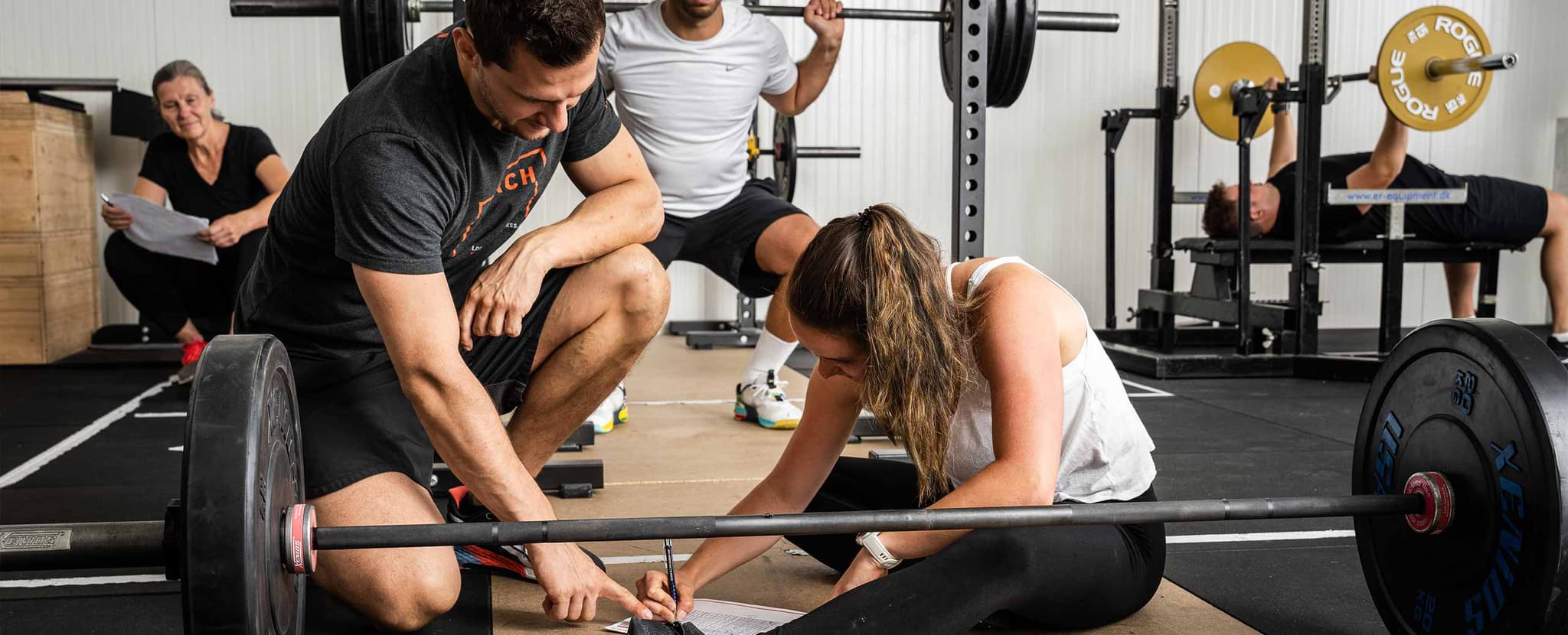 Trainer helps client with barbell training A trainer kneels to help a client adjust a barbell in a busy gym.