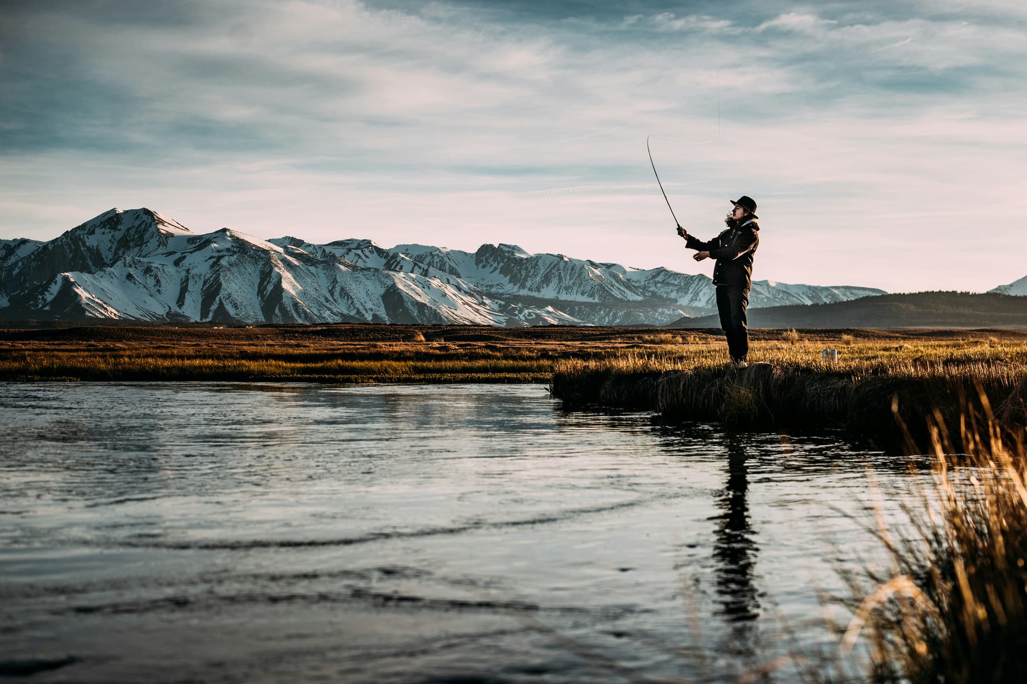 Fly fisherman casting in a mountain river A fly fisherman casts his line into a river, with snow-capped mountains and tundra landscape in the background.