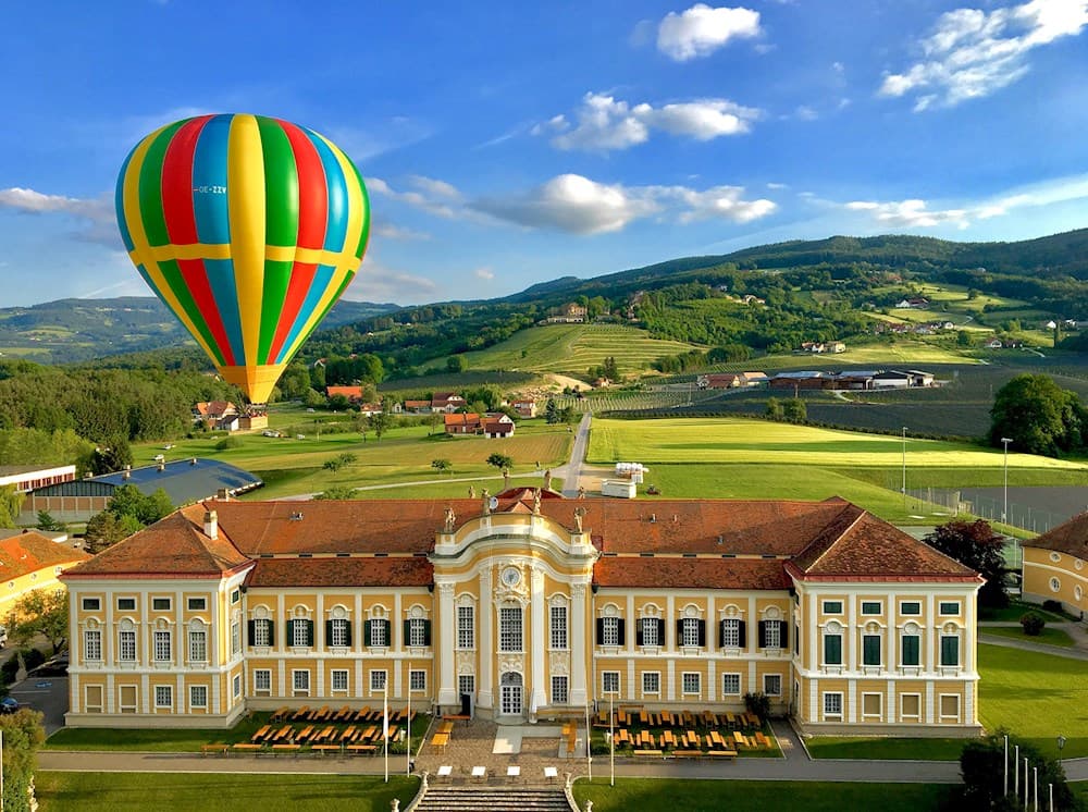 Hot Air Balloon Over Baroque Castle A colorful hot air balloon flies over a baroque castle with green hills in the background.