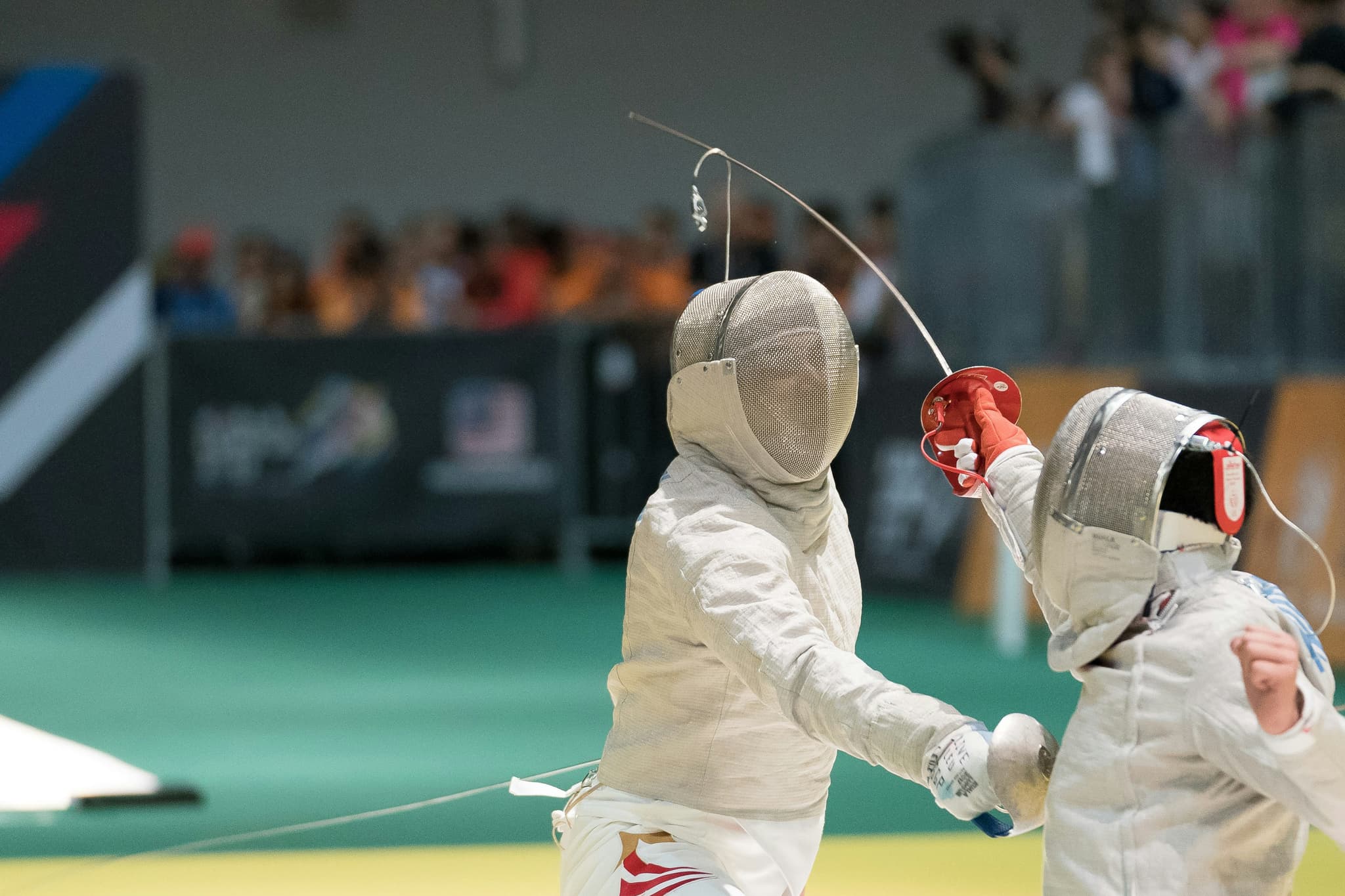 Fencers Competing in Indoor Arena Two fencers in white protective gear and masks competing on a green mat during an indoor fencing competition with spectators in the background.