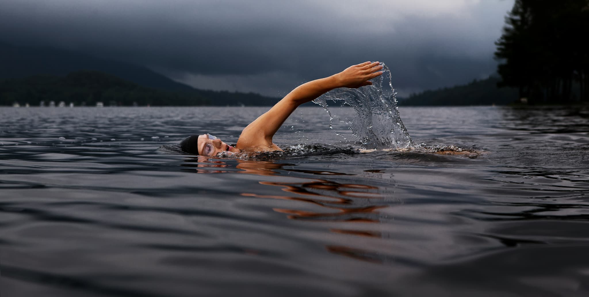 Swimmer in Mountain Lake Swimmer performing freestyle swimming in a calm mountain lake, with forested hills and cloudy sky in the background.