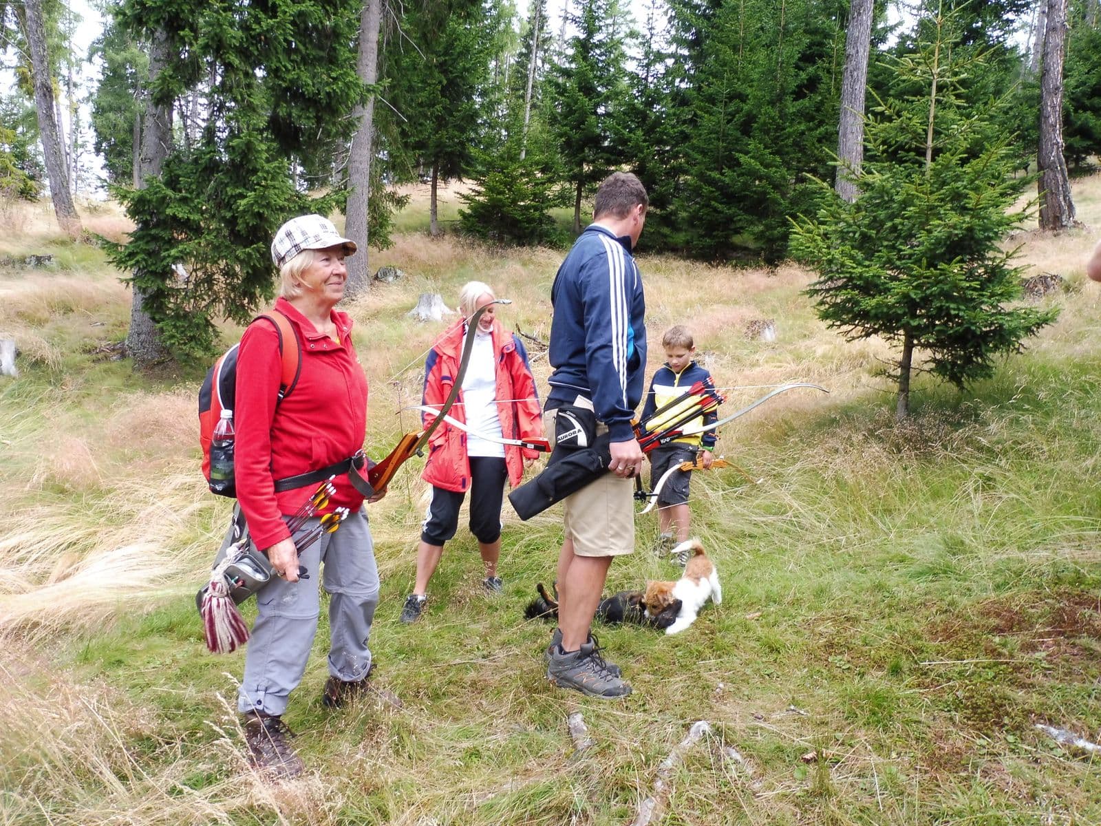 Bogenschießen mit der Familie im Wald Eine Familiengruppe mit Bögen und Pfeilen steht zusammen in einem bewaldeten Außenbereich mit hohen Bäumen und grünem Gelände.