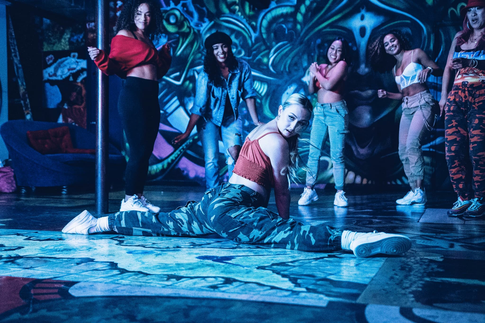 Dance group performs under blue stage lighting A dancer performs a split on the floor while a group dances in the background under blue lighting.