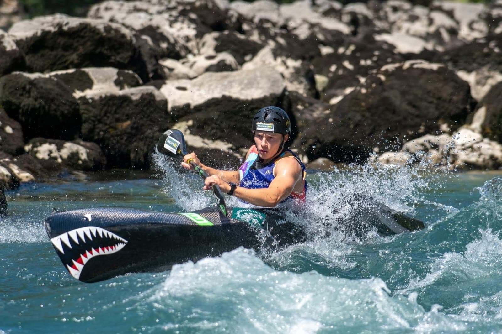 Kayaker paddling through whitewater rapids Kayaker wearing a helmet paddling through splashing whitewater near a rocky shore.