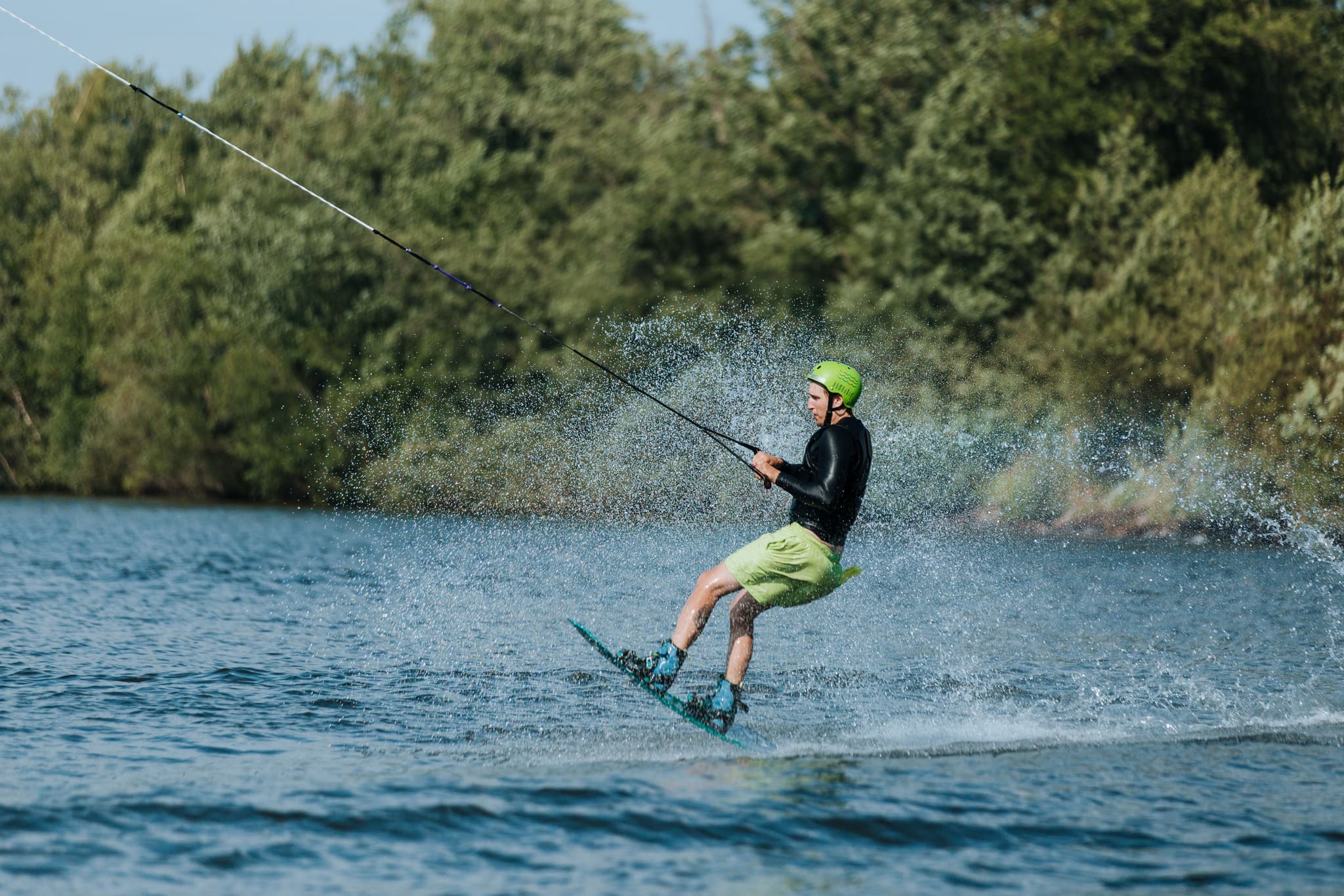 Wakeboarding on Lake With Forest Person wakeboarding on a lake while being towed by a rope, wearing a green helmet and life vest, with dense forest in the background.