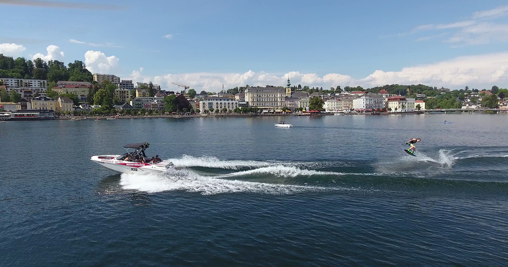 Wakeboarder on the lake with harbor city A wakeboarder is being towed behind a boat across a lake, with a harbor city and green hills in the background.