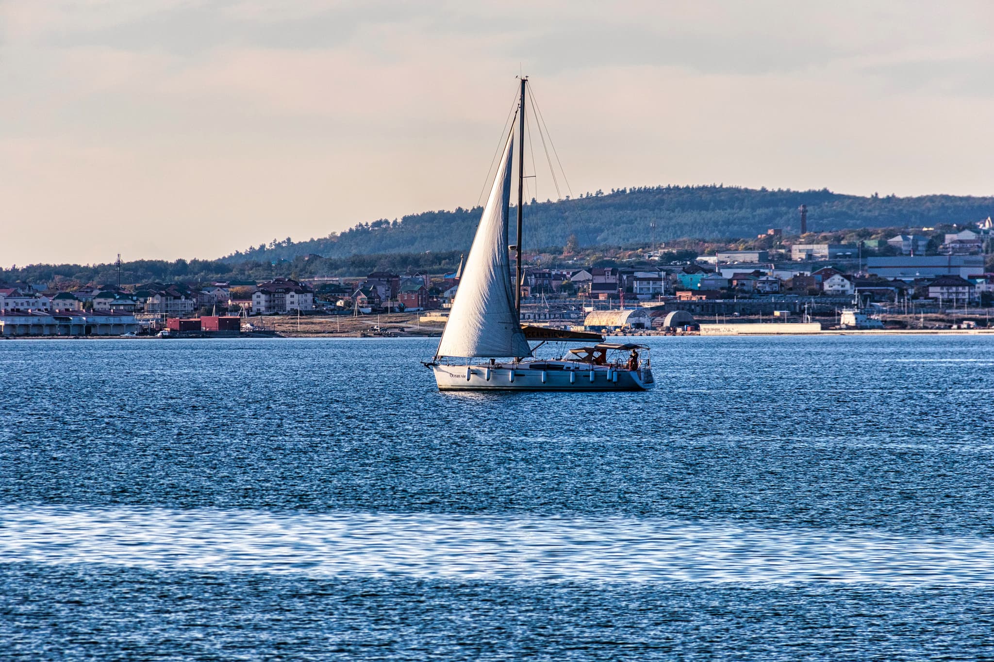 Sailboat on Coastal Waters Sailboat with white sail on blue water, residential waterfront and forested hills in background.