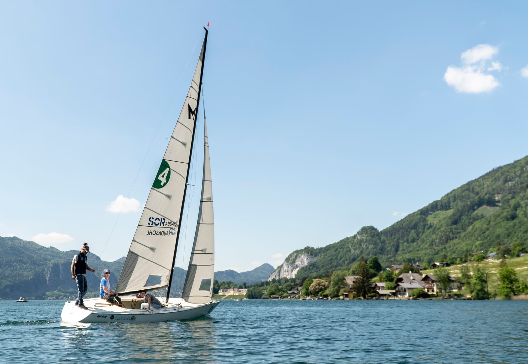 Segelboot auf Alpensee mit Bergen Segelboot mit weißen Segeln auf einem Alpensee, umgeben von bewaldeten Bergen und Häusern am Seeufer.