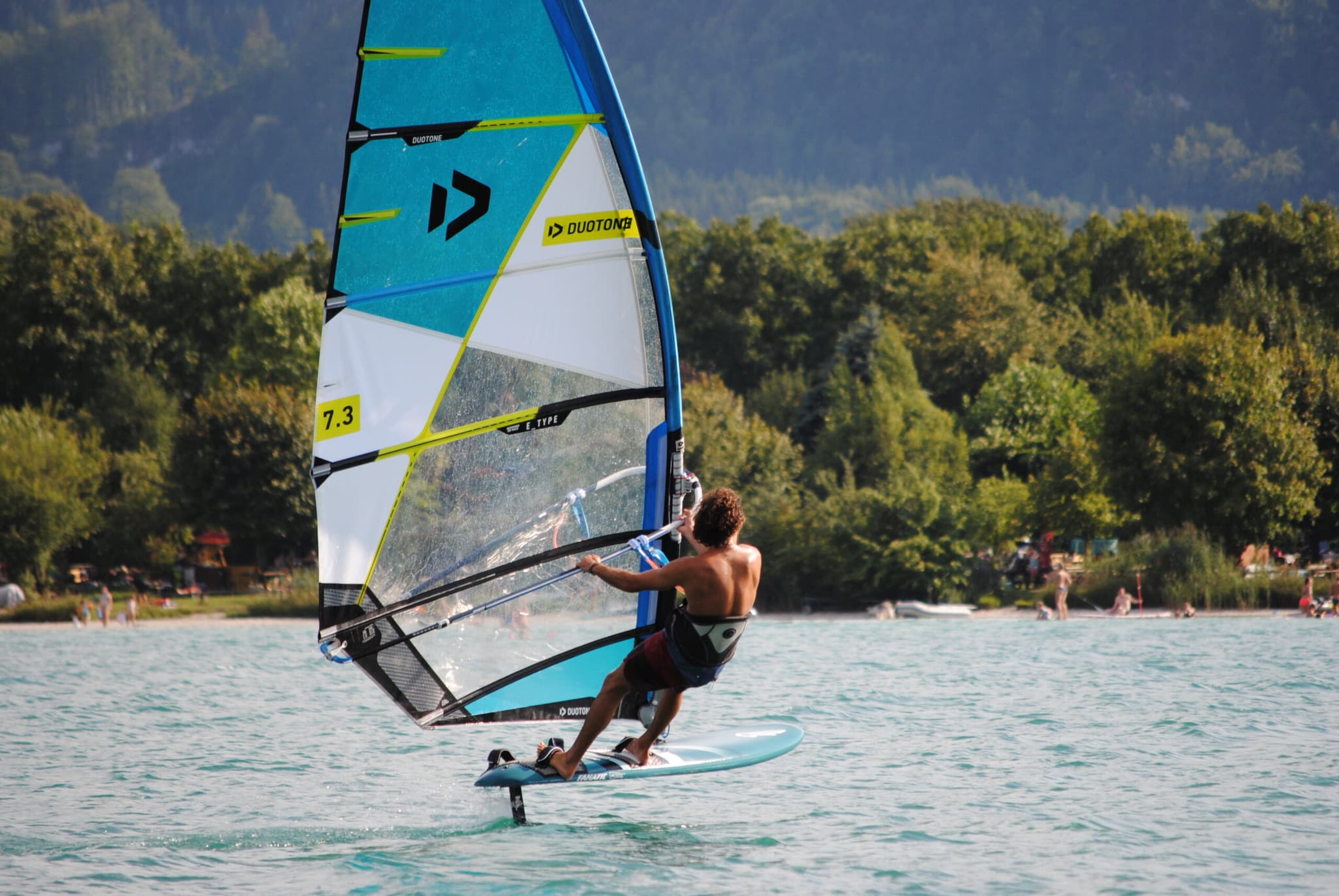 Windsurfer auf Alpensee Windsurfer auf einem Foilboard mit türkis-weißem Segel auf einem Alpensee, bewaldete Berge im Hintergrund.