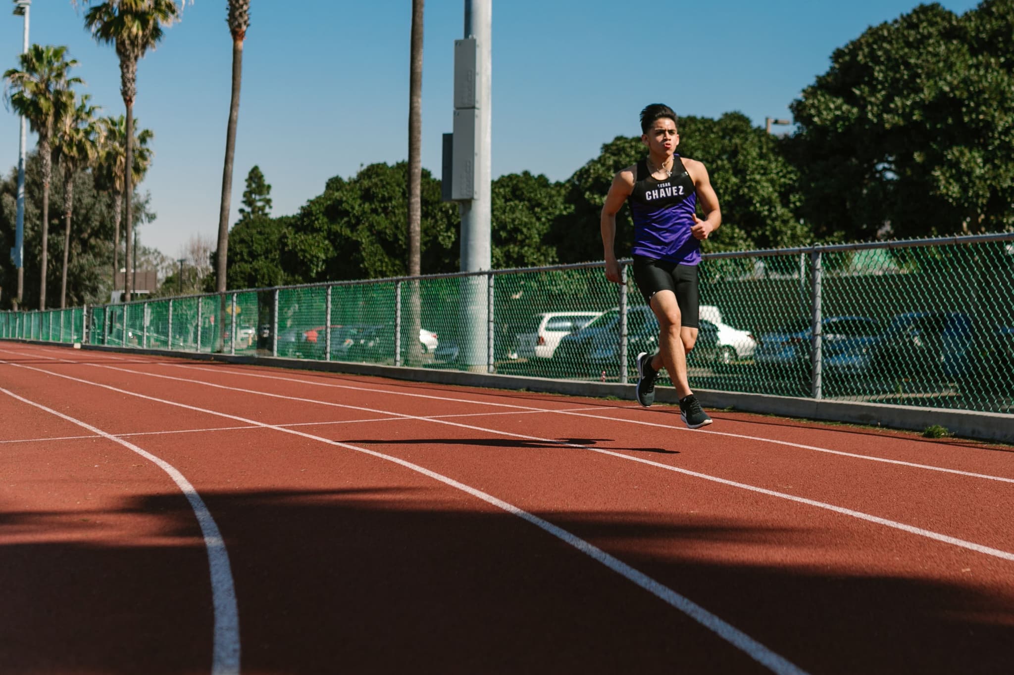 Runner on Outdoor Track Athlete running on a red outdoor track with palm trees and green fencing in the background.