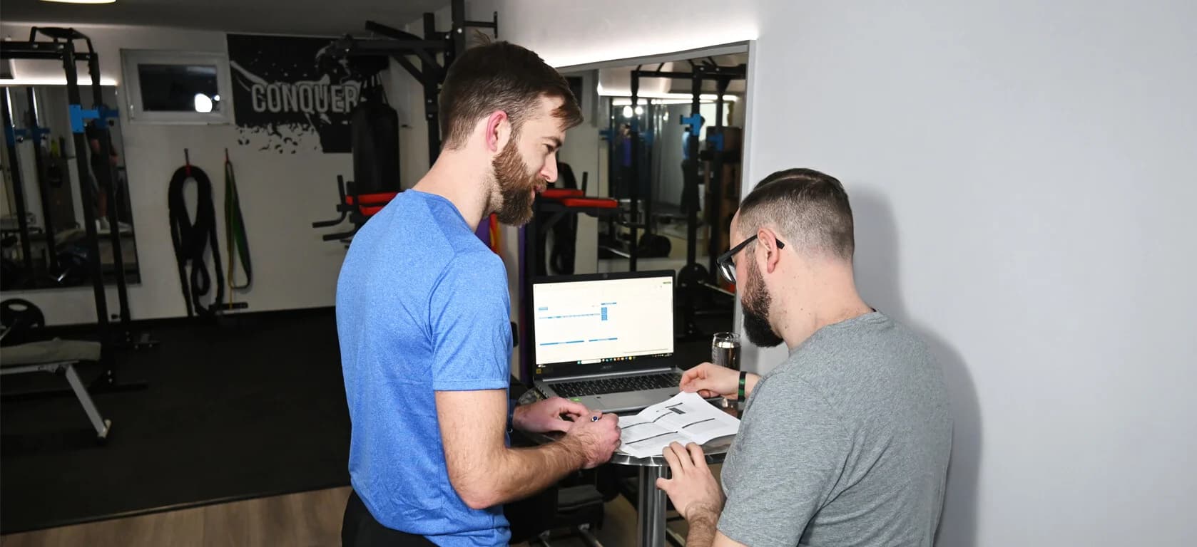 Reviewing Documents in a Home Gym Two men discussing paperwork near a laptop in a home gym.