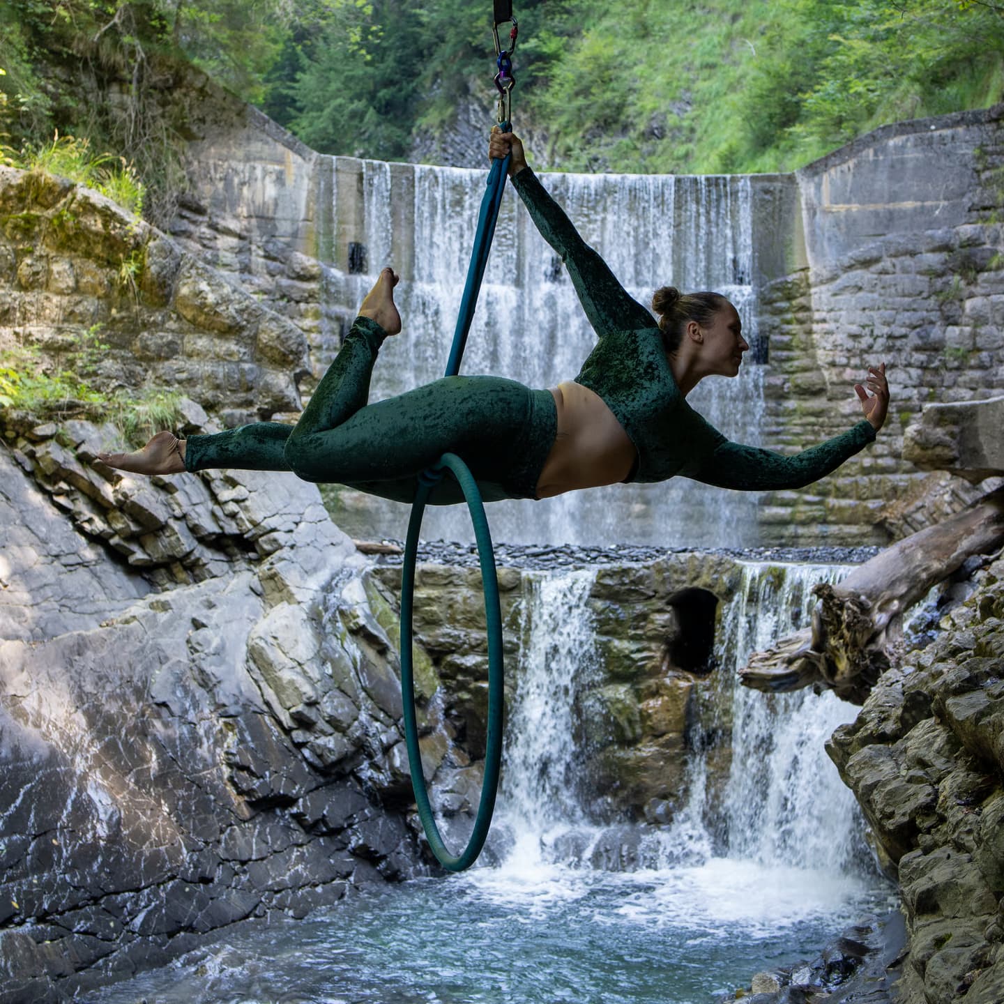 Aerial Hoop Performance Over a Waterfall Aerial hoop artist performing above a rocky waterfall in a green, forested canyon.