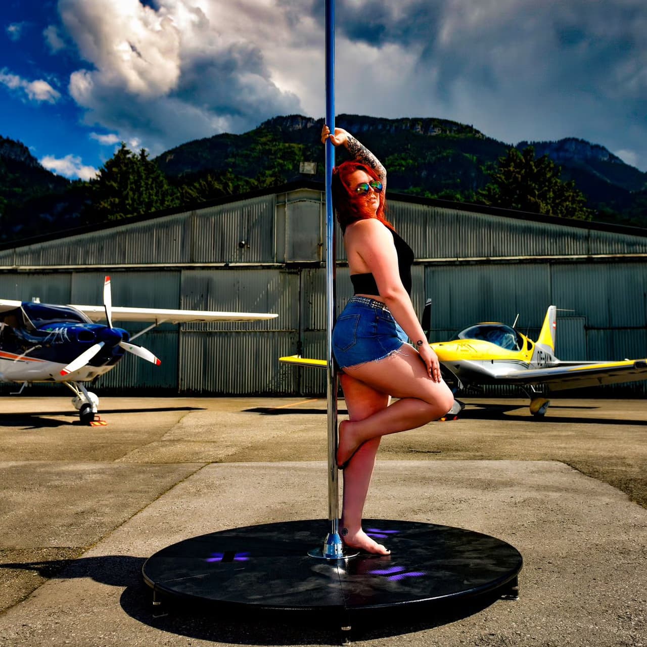 Pole Dancer at Mountain Airfield Person posing on a dance pole near small planes at an airfield with mountains behind.