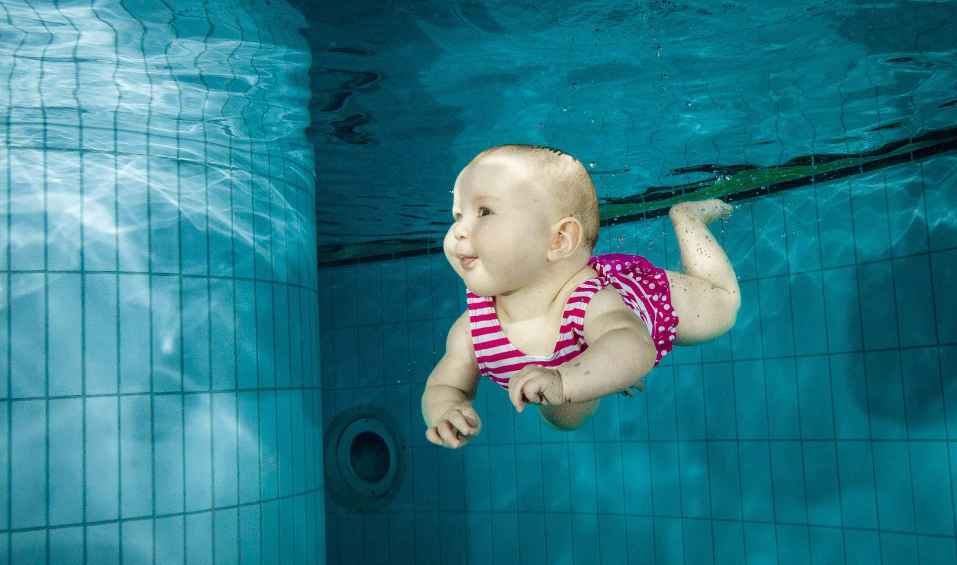 Säugling schwimmt unter Wasser im Becken Säugling schwimmt unter Wasser in einem Becken und trägt einen rosa-weiß gestreiften Badeanzug.