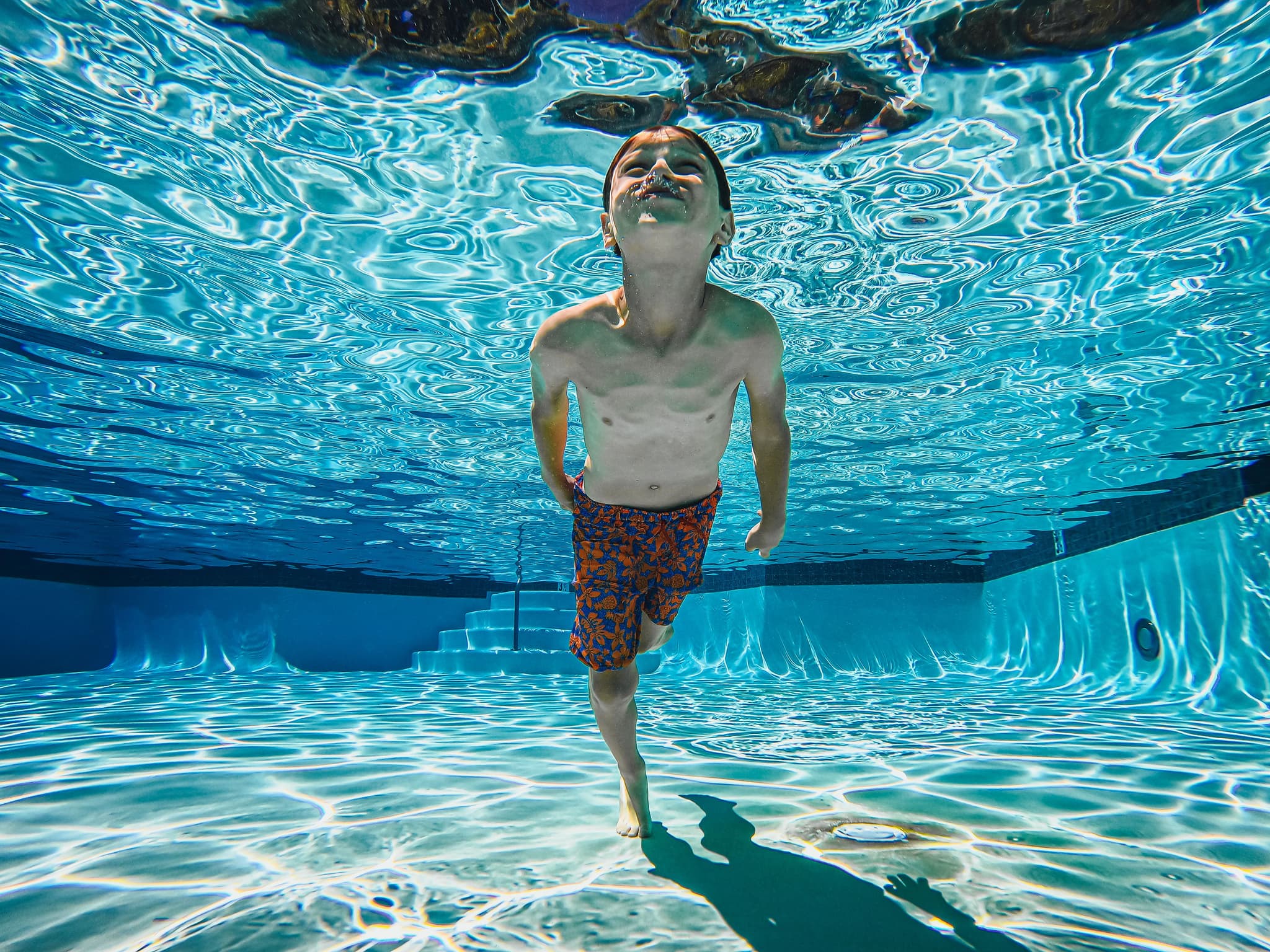 Child swimming underwater in the pool Child swimming underwater in a clear pool, looking upward, wave patterns visible above and below the water.
