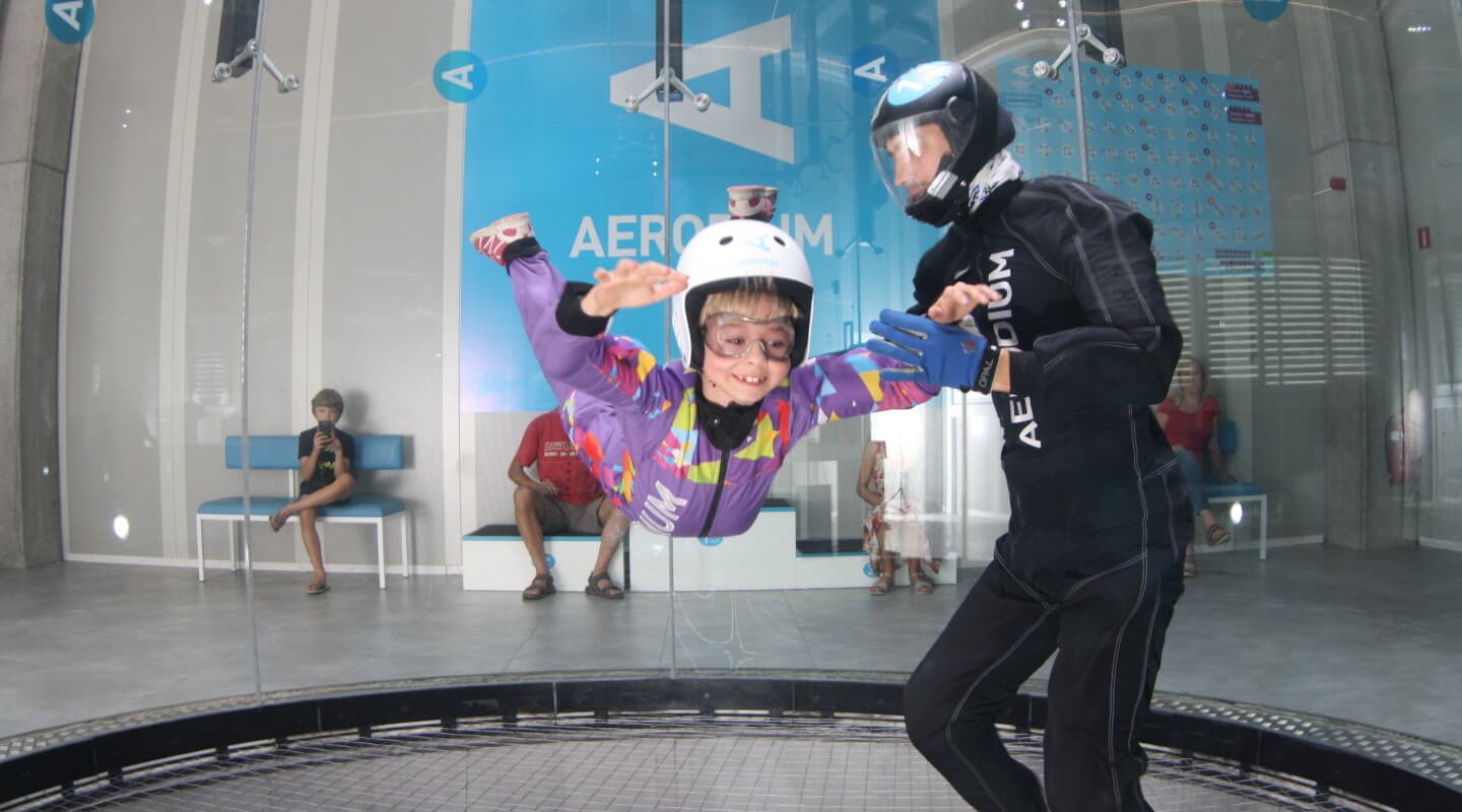 Child Indoor Skydiving with Instructor Child in purple flight suit and white helmet flying indoors with instructor in black suit spotting nearby.