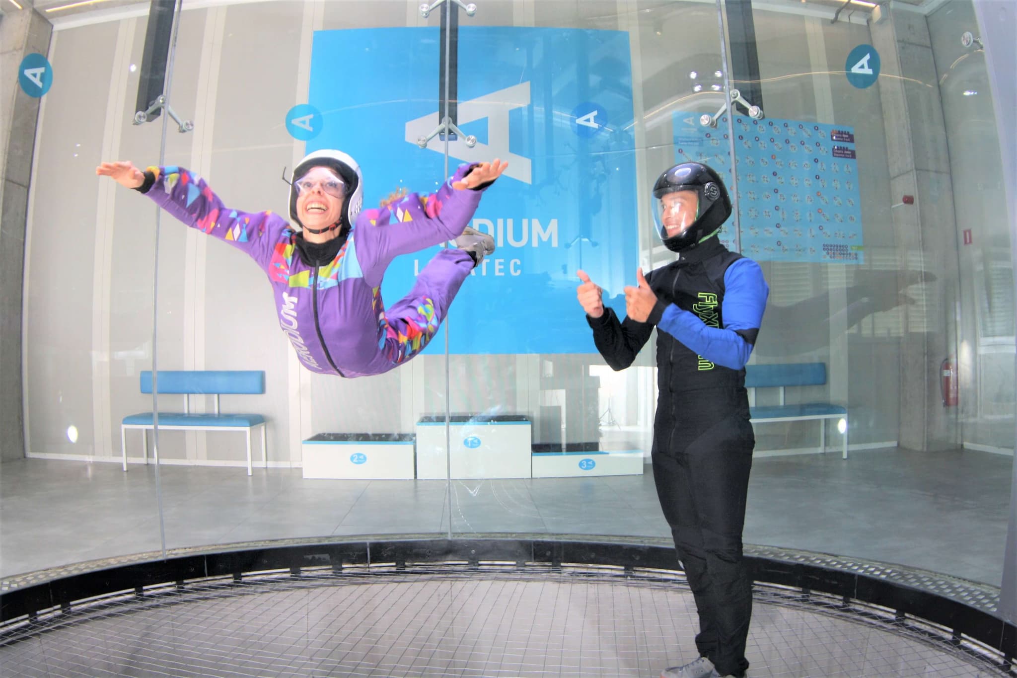 Indoor Skydiving Wind Tunnel Experience Two people in an indoor skydiving wind tunnel, one in a purple jumpsuit with arms spread wide, the other in black and blue gear standing on the padded floor.