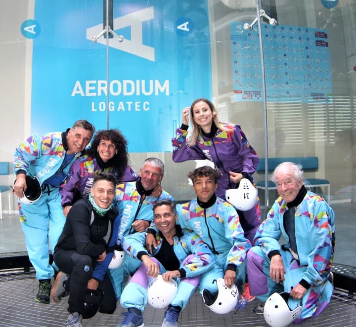 Skydiving Team at Indoor Facility Group of skydivers in colorful jumpsuits posing together at an indoor skydiving facility with Aerodium Logatec signage.