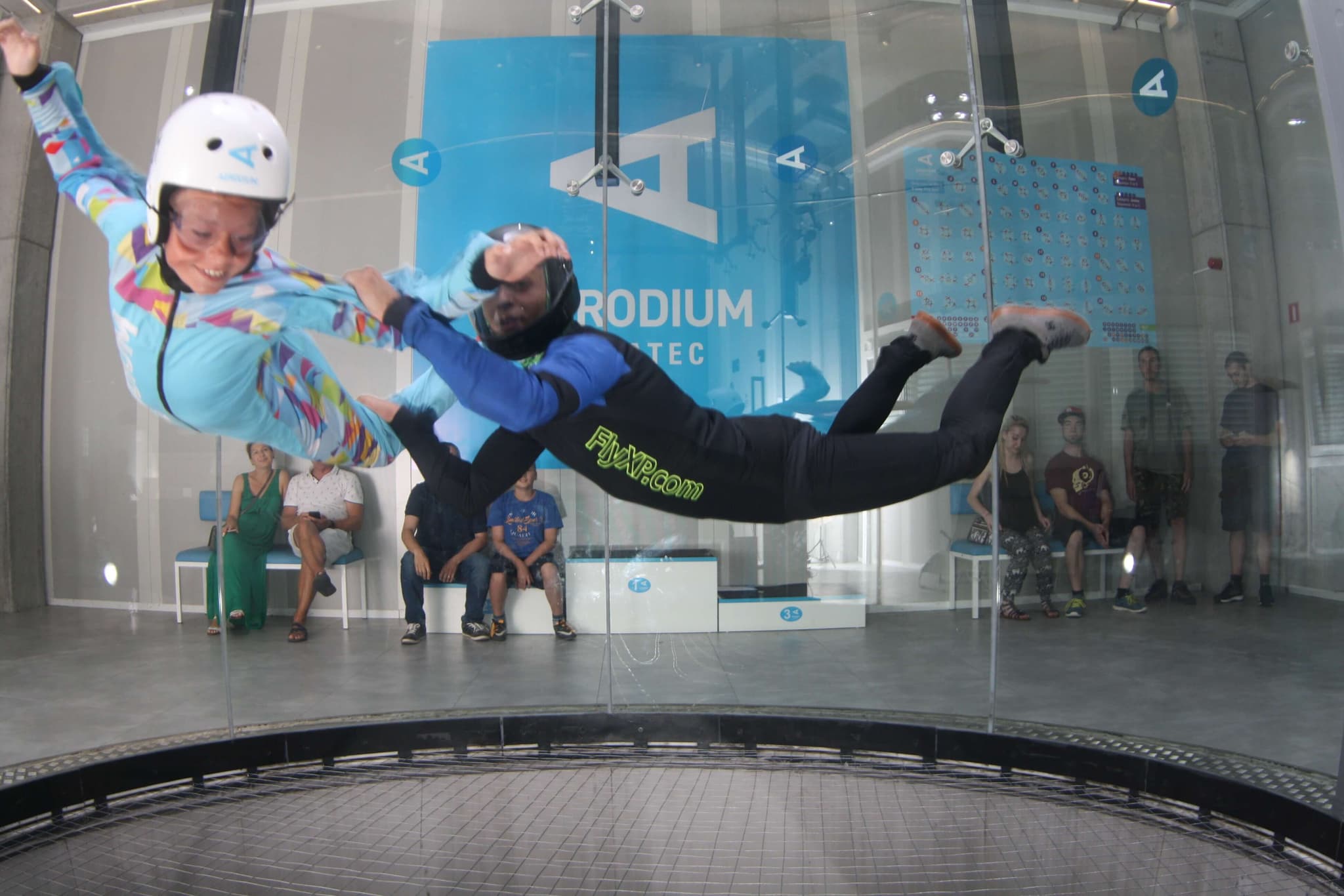Indoor Skydiving Wind Tunnel Experience Two people in helmets and jumpsuits flying horizontally in an indoor skydiving wind tunnel with spectators watching from the sides.